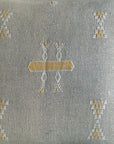 A close-up of a woven throw pillow in a textured gray fabric, featuring a central embroidered geometric motif in white and mustard yellow, with matching motifs in the corners.