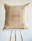 A square pale orange embroidered geometric patterns, resting on a white and light wood tripod stool against a white textured wall.