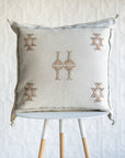 A square off-white throw pillow with embroidered geometric patterns in brown and beige, displayed on a white and light wood stool against a textured white wall.