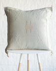 A square decorative throw pillow in a light tan linen color with embroidered geometric patterns, displayed on a white table with wooden legs against a white textured wall.