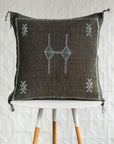 A square dark brown woven throw pillow with embroidered geometric patterns in white and teal, displayed on a white table with wooden legs against a white textured wall.