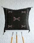 A square dark brown woven throw pillow with geometric embroidered patterns, displayed on a white and light wood stool against a white textured wall.