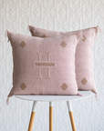 Two dusty pink textured throw pillows with embroidered details, displayed on a white and light wood side table against a white patterned wall.