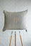 A square woven throw pillow in a light gray linen fabric with a central geometric pattern in mustard yellow and white, displayed on a white and wood stool against a textured white wall.