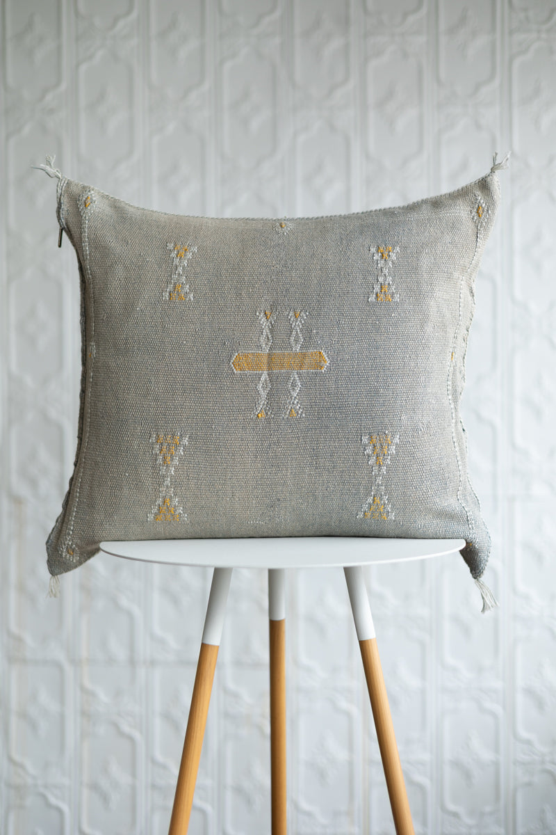 A square woven throw pillow in a light gray linen fabric with a central geometric pattern in mustard yellow and white, displayed on a white and wood stool against a textured white wall.