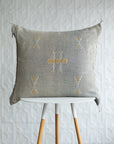 A square woven throw pillow in a light gray linen fabric with a central geometric pattern in mustard yellow and white, displayed on a white and wood stool against a textured white wall.