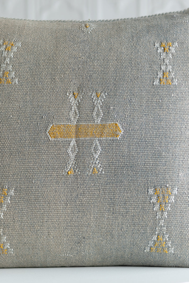 A close-up of a woven throw pillow in a textured gray fabric, featuring a central embroidered geometric motif in white and mustard yellow, with matching motifs in the corners.