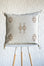 A square off-white throw pillow with embroidered geometric patterns in brown and beige, displayed on a white and light wood stool against a textured white wall.