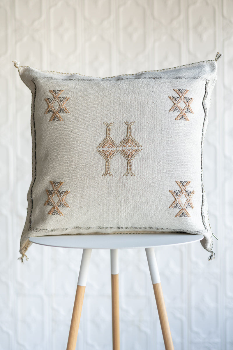 A square off-white throw pillow with embroidered geometric patterns in brown and beige, displayed on a white and light wood stool against a textured white wall.