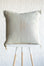 A square decorative throw pillow in a light tan linen color with embroidered geometric patterns, displayed on a white table with wooden legs against a white textured wall.