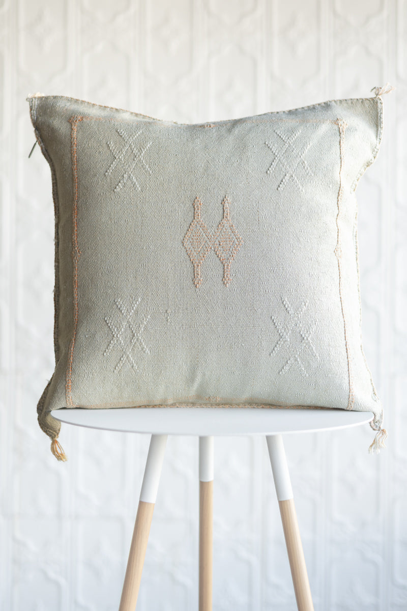 A square decorative throw pillow in a light tan linen color with embroidered geometric patterns, displayed on a white table with wooden legs against a white textured wall.
