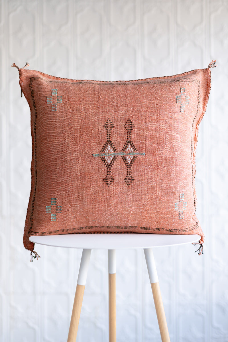 A square woven throw pillow in a dusty terracotta pink color with a central geometric diamond pattern and small cross motifs in the corners, displayed on a white and light wood stool against a white textured wall.