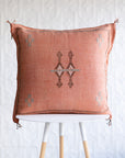 A square woven throw pillow in a dusty terracotta pink color with a central geometric diamond pattern and small cross motifs in the corners, displayed on a white and light wood stool against a white textured wall.