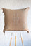 A square light orange woven throw pillow with a central geometric embroidered pattern and corner diamond motifs, displayed on a white and light wood stool against a white textured wall.