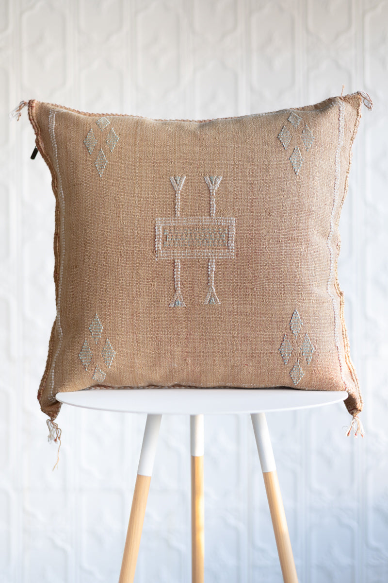A square light orange woven throw pillow with a central geometric embroidered pattern and corner diamond motifs, displayed on a white and light wood stool against a white textured wall.
