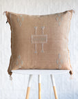 A square light orange woven throw pillow with a central geometric embroidered pattern and corner diamond motifs, displayed on a white and light wood stool against a white textured wall.