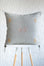 A square light gray throw pillow with embroidered geometric diamond patterns in white, dusty pink, and ochre yellow, displayed on a white stool against a white textured wall.