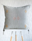 A square light gray throw pillow with embroidered geometric diamond patterns in white, dusty pink, and ochre yellow, displayed on a white stool against a white textured wall.