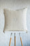 A square off-white woven throw pillow with light blue geometric embroidery, displayed on a white and light wood stool against a textured white wall.