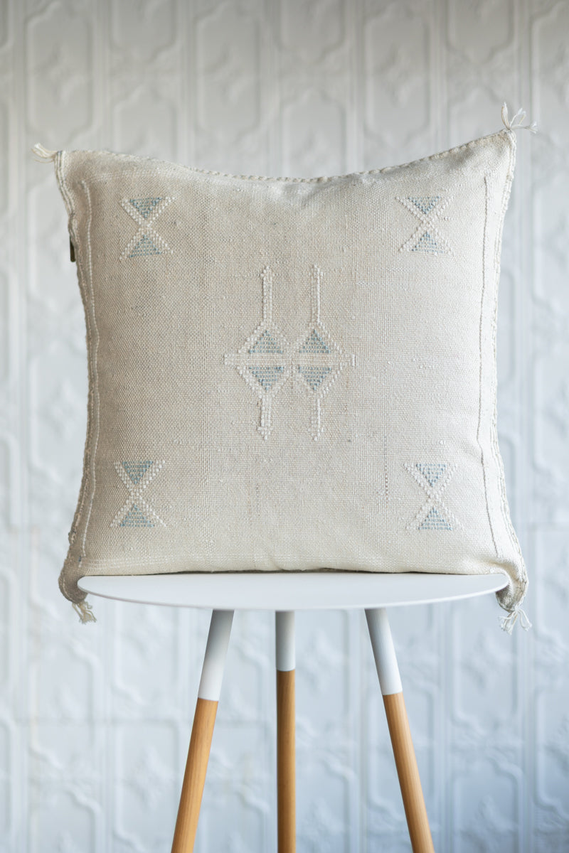 A square off-white woven throw pillow with light blue geometric embroidery, displayed on a white and light wood stool against a textured white wall.