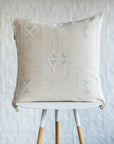 A square off-white woven throw pillow with light blue geometric embroidery, displayed on a white and light wood stool against a textured white wall.