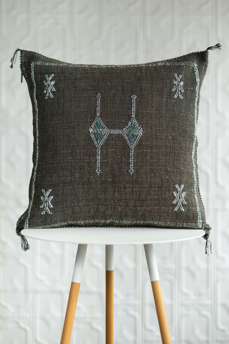 A square dark brown woven throw pillow with embroidered geometric patterns in white and teal, displayed on a white table with wooden legs against a white textured wall.