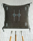 A square dark brown woven throw pillow with embroidered geometric patterns in white and teal, displayed on a white table with wooden legs against a white textured wall.