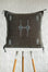 A square dark brown woven throw pillow with embroidered geometric patterns in white and teal, displayed on a white table with wooden legs against a white textured wall.