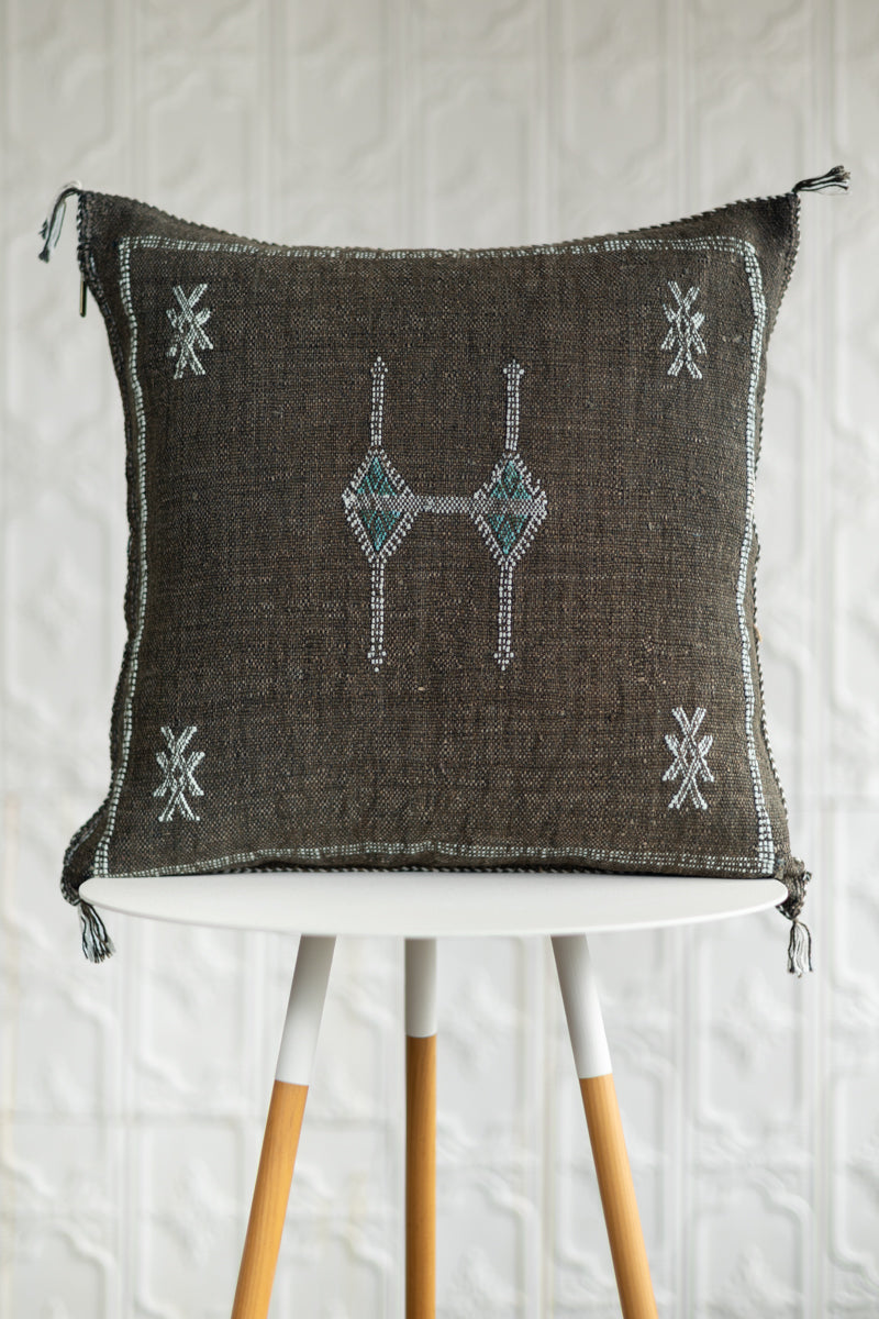 A square dark brown woven throw pillow with embroidered geometric patterns in white and teal, displayed on a white table with wooden legs against a white textured wall.