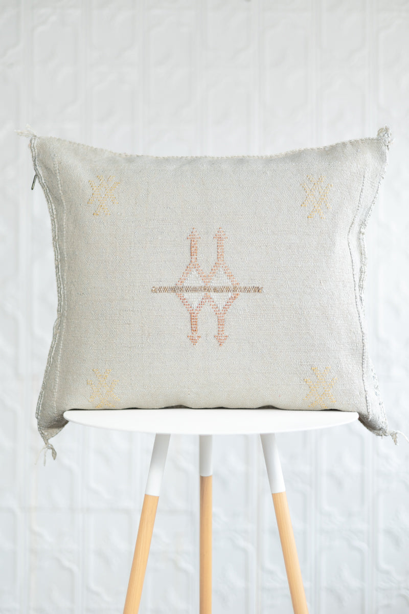 A square beige linen throw pillow with a central embroidered geometric design in muted orange and gold, resting on a white table with light wood legs against a white textured wall.