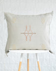A square beige linen throw pillow with a central embroidered geometric design in muted orange and gold, resting on a white table with light wood legs against a white textured wall.