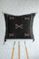 A square dark brown woven throw pillow with geometric embroidered patterns, displayed on a white and light wood stool against a white textured wall.