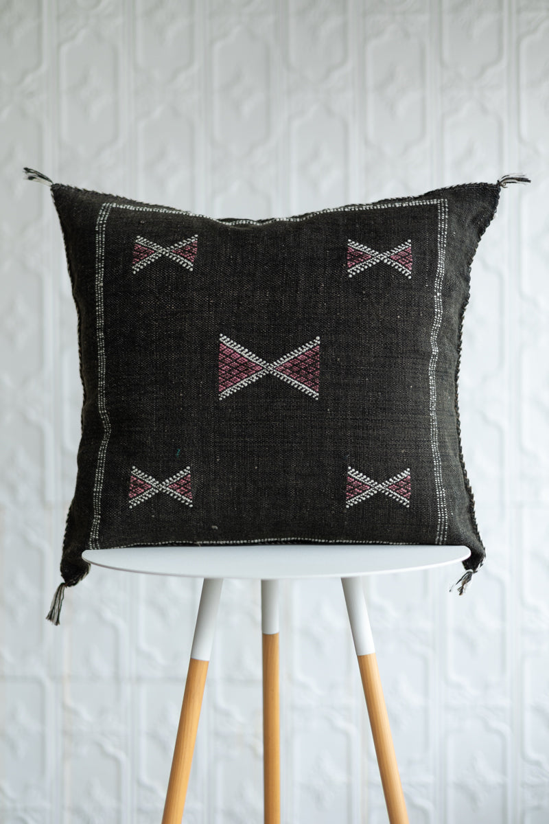 A square dark brown woven throw pillow with geometric embroidered patterns, displayed on a white and light wood stool against a white textured wall.