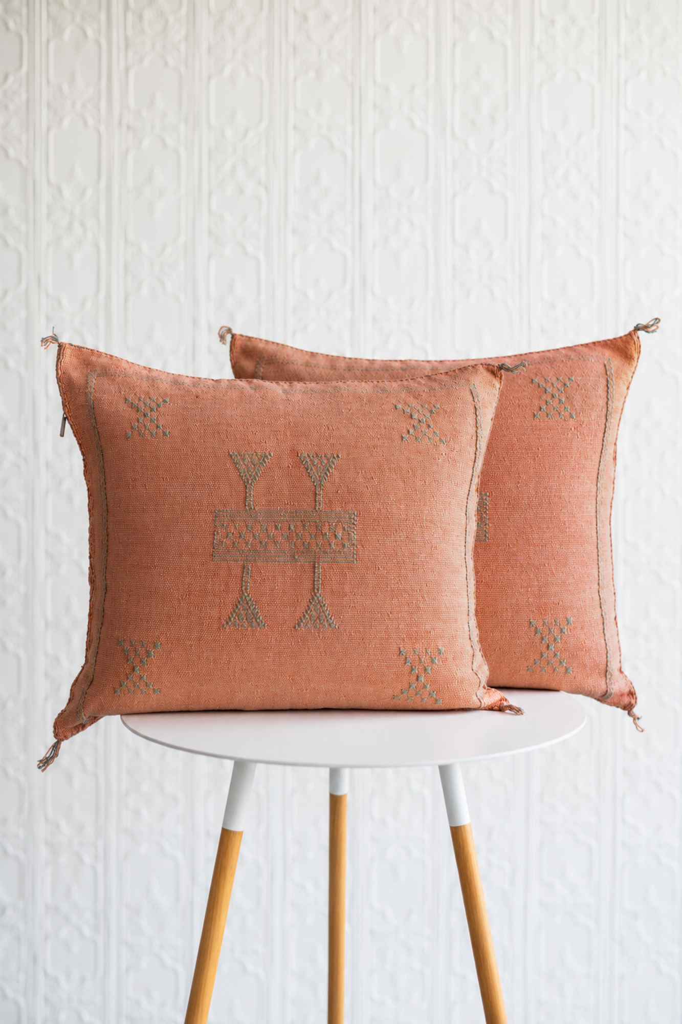 Two square terracotta throw pillows with embroidered geometric patterns, displayed on a white table with wooden legs against a white textured wall.