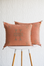 Two square terracotta throw pillows with embroidered geometric patterns, displayed on a white table with wooden legs against a white textured wall.